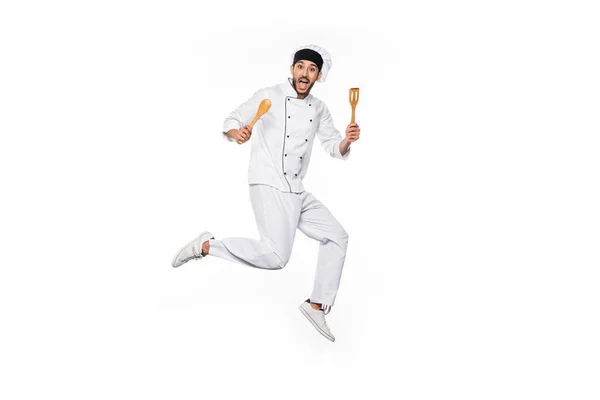 Excited chef in hat and uniform jumping and holding wooden spatula and spoon isolated on white — Stock Photo