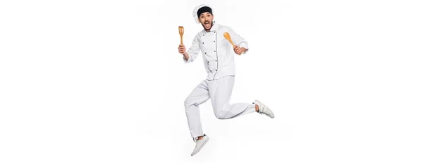 Excited chef in hat and uniform jumping and holding wooden spatula and spoon isolated on white, banner — Stock Photo