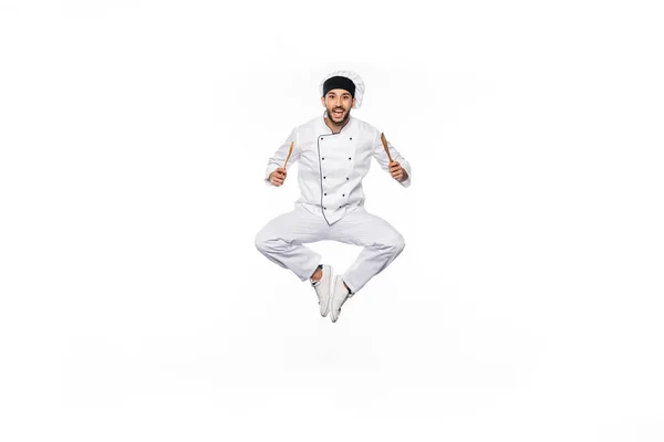 Chef alegre en sombrero y uniforme saltando y sosteniendo espátula de madera y cuchara aislada en blanco - foto de stock