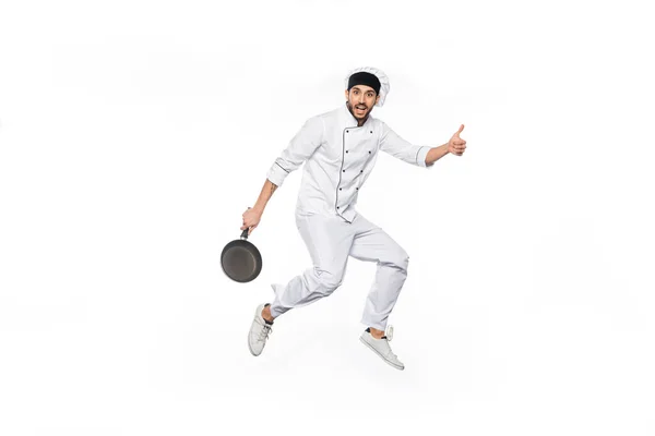 Cheerful chef in hat and uniform jumping, showing thumb up while holding frying pan isolated on white — Stock Photo