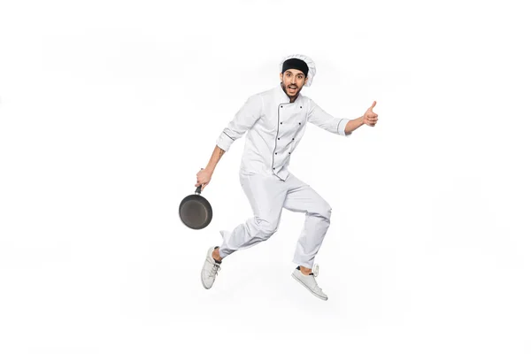 Alegre chef en sombrero y uniforme saltando, mostrando el pulgar hacia arriba mientras sostiene la sartén aislada en blanco - foto de stock