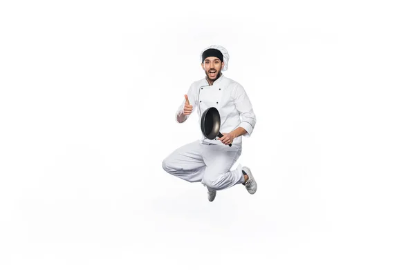 Alegre chef con sombrero y uniforme saltando, mostrando el pulgar hacia arriba mientras sostiene la sartén aislada en blanco - foto de stock