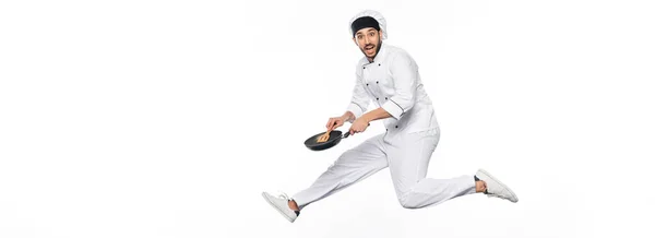 Cheerful chef in hat and uniform jumping and holding frying pan with wooden spatula isolated on white, banner — Stock Photo