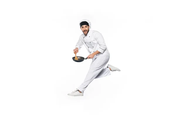 Chef sonriente en sombrero y uniforme saltando y sosteniendo sartén con espátula de madera aislada en blanco - foto de stock