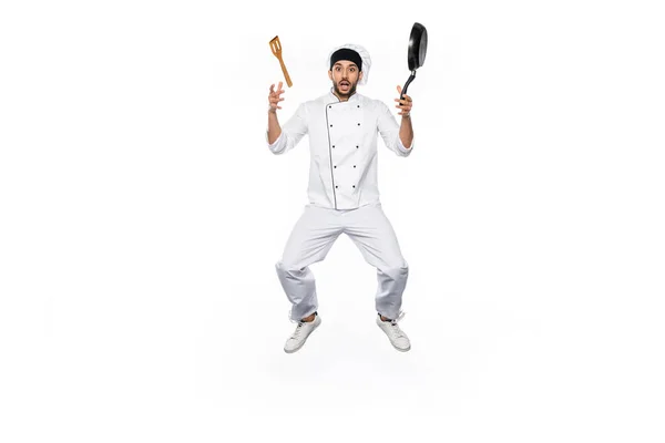 Chef chocado em chapéu e levitação uniforme perto de frigideira voadora com espátula de madeira isolada em branco — Fotografia de Stock