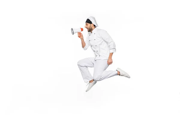 Side view of young chef in hat and uniform levitating and speaking in megaphone isolated on white — Stock Photo