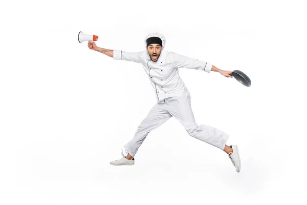 Überraschter junger Koch mit Hut und Uniform schwebt, während er Bratpfanne und Megafon isoliert auf weiß hält — Stockfoto