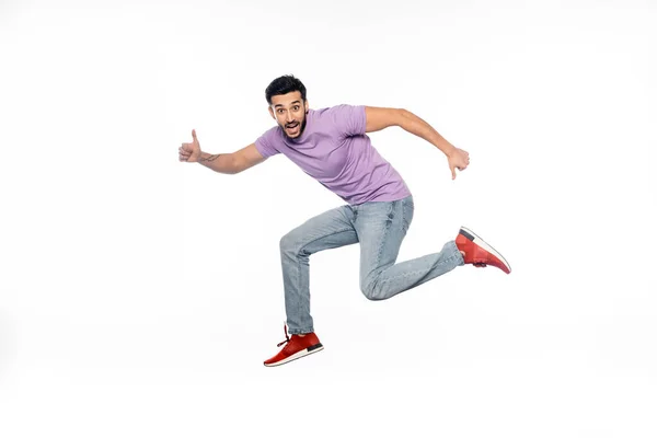 Hombre sorprendido en jeans y camiseta púrpura levitando mientras muestra los pulgares hacia arriba aislado en blanco - foto de stock