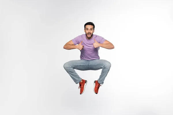 Hombre sorprendido en jeans y camiseta púrpura levitando mientras muestra los pulgares hacia arriba en blanco - foto de stock