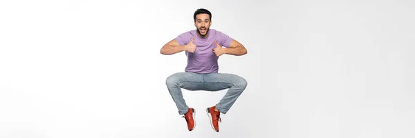 Hombre sorprendido en jeans y camiseta púrpura levitando mientras muestra los pulgares hacia arriba en blanco, bandera - foto de stock