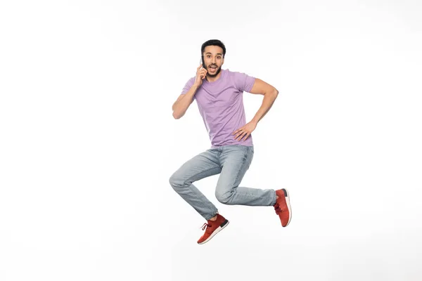 Изумленный человек в джинсах и фиолетовой футболке левитирует, разговаривая по мобильному телефону на белом — стоковое фото