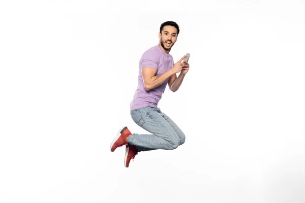 Hombre sorprendido en jeans y camiseta púrpura levitando mientras sostiene el teléfono inteligente en blanco - foto de stock