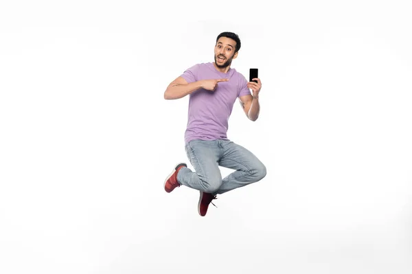 Amazed man in jeans and purple t-shirt levitating while pointing at smartphone on white — Stock Photo