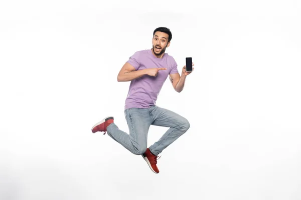Excited man in jeans and purple t-shirt levitating while pointing at smartphone on white — Stock Photo