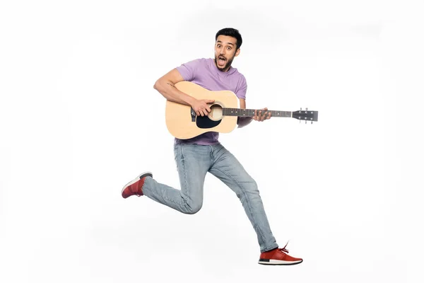 Изумленный человек в джинсах и фиолетовой футболке, левитирующий во время игры на акустической гитаре на белом — стоковое фото