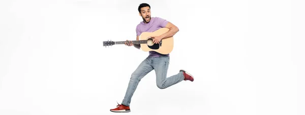 Homme étonné en jeans et t-shirt violet lévitant tout en jouant de la guitare acoustique sur blanc, bannière — Photo de stock