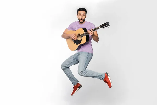 Shocked man in jeans and purple t-shirt levitating while playing acoustic guitar on white — Stock Photo