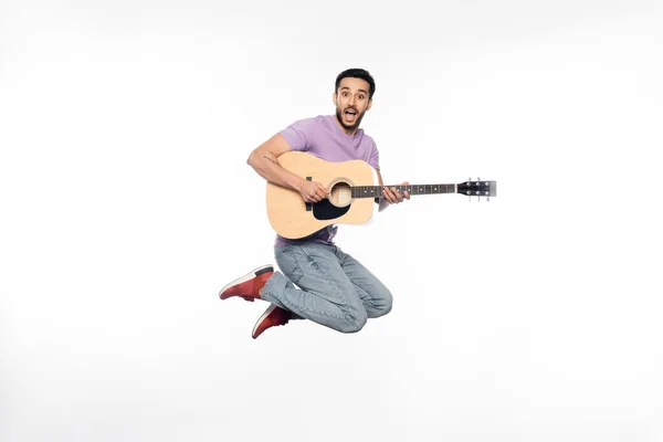 Homem positivo em jeans e camiseta roxa levitando enquanto tocava guitarra acústica no branco — Fotografia de Stock