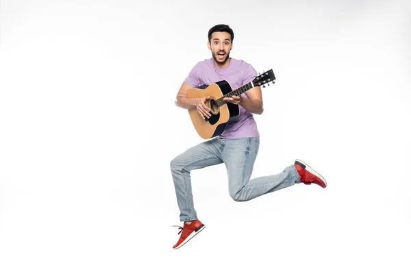 Hombre sorprendido en pantalones vaqueros azules y camiseta púrpura levitando mientras toca la guitarra acústica en blanco - foto de stock