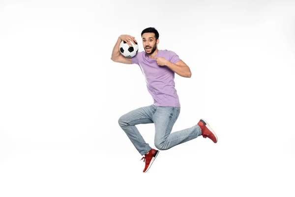 Hombre feliz en jeans y camiseta púrpura levitando mientras señala el fútbol en blanco - foto de stock