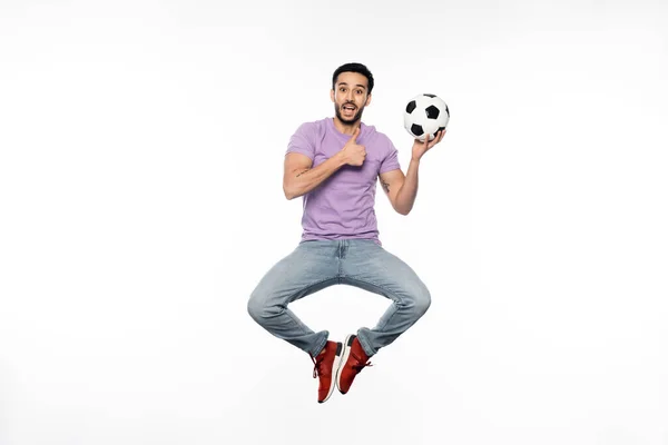 Hombre sorprendido en jeans y camiseta púrpura levitando mientras sostiene el fútbol y mostrando el pulgar hacia arriba en blanco - foto de stock