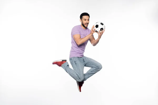 Happy man in jeans and purple t-shirt levitating while looking at football on white — Stock Photo