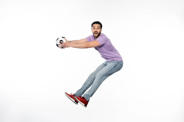 Aufgeregter Mann in Jeans und lila T-Shirt schwebt mit Fußball auf weißem Grund — Stockfoto