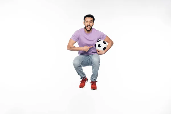 Amazed man in jeans and purple t-shirt levitating while pointing at football on white — Stock Photo
