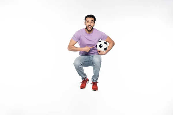 Homem maravilhado em jeans e camiseta roxa levitando enquanto apontava para o futebol em branco — Fotografia de Stock