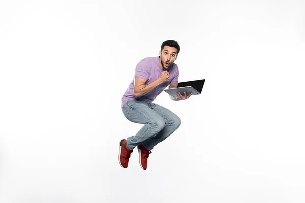 Hombre sorprendido en jeans y camiseta púrpura levitando mientras sostiene el ordenador portátil en blanco - foto de stock