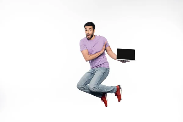 Hombre sorprendido en jeans y camiseta púrpura levitando mientras sostiene el ordenador portátil en blanco - foto de stock