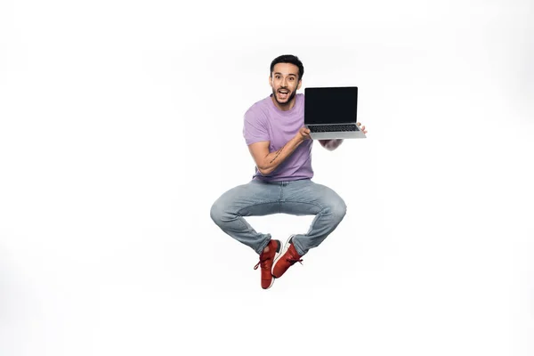 Hombre positivo en jeans y camiseta púrpura levitando mientras sostiene el ordenador portátil con pantalla en blanco - foto de stock