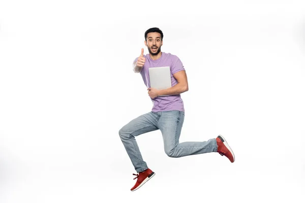 Positive man in jeans and purple t-shirt levitating while holding laptop and showing thumb up on white — Stock Photo