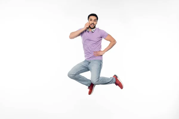 Homem positivo em fones de ouvido sem fio falando no celular e levitando no branco — Fotografia de Stock