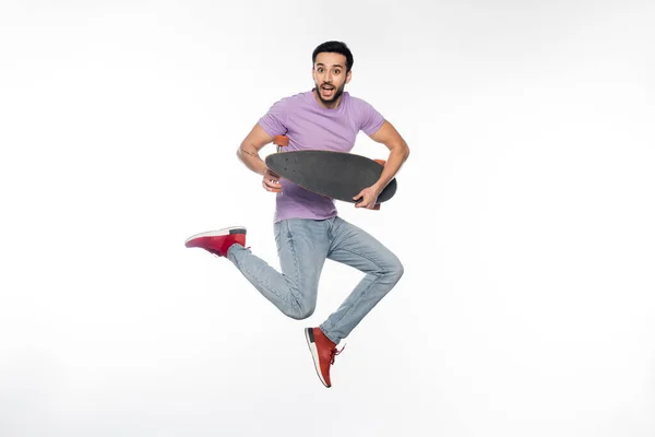 Uomo positivo in jeans e t-shirt viola che salta con longboard su bianco — Foto stock