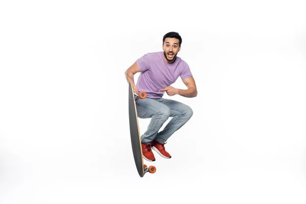 Positive man in jeans and purple t-shirt jumping and pointing at longboard on white — Stock Photo