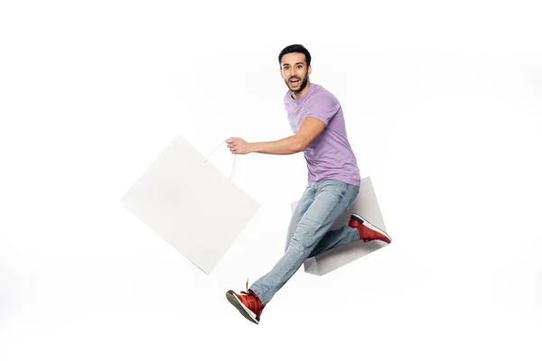 Homem alegre em jeans e camiseta roxa levitando com sacos de compras isolados em branco — Fotografia de Stock