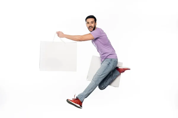 Felice uomo in jeans e t-shirt viola levitante con shopping bags isolato su bianco — Foto stock