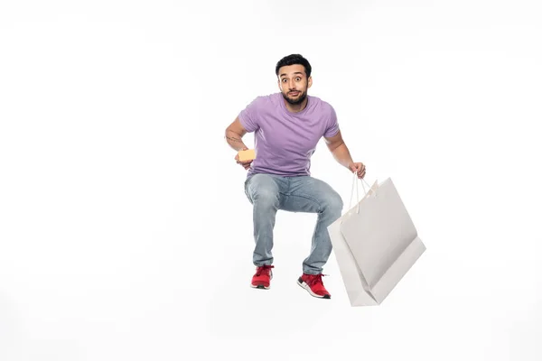 Hombre en jeans y camiseta púrpura levitando mientras sostiene la tarjeta de crédito y el bolso de compras en blanco - foto de stock