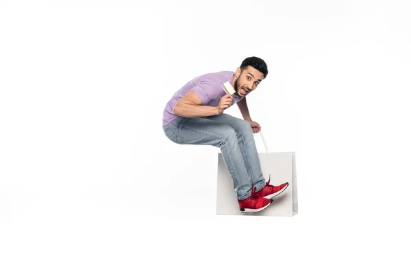 Hombre sorprendido en jeans y camiseta púrpura levitando mientras sostiene la tarjeta de crédito y el bolso de compras en blanco - foto de stock