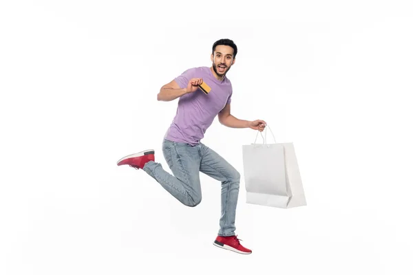 Hombre excitado en jeans y camiseta púrpura levitando mientras sostiene la tarjeta de crédito y el bolso de compras en blanco - foto de stock