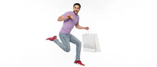 Uomo eccitato in jeans e t-shirt viola levitante mentre tiene carta di credito e shopping bag su bianco, banner — Foto stock