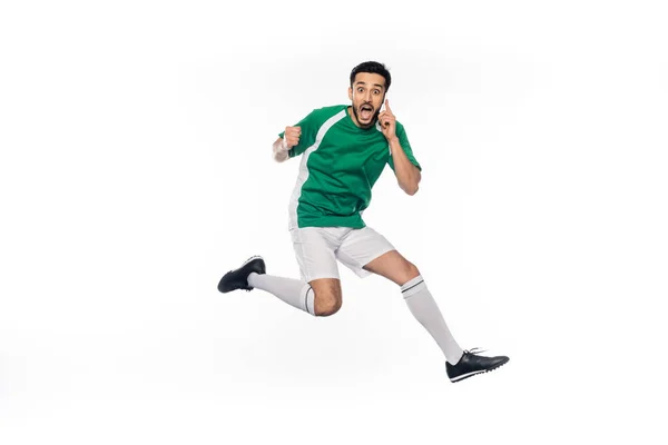 Giocatore di calcio stupito in uniforme che salta mentre parla su smartphone su bianco — Foto stock