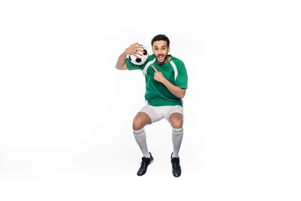 Amazed football player in uniform jumping while pointing at soccer ball isolated on white — Stock Photo