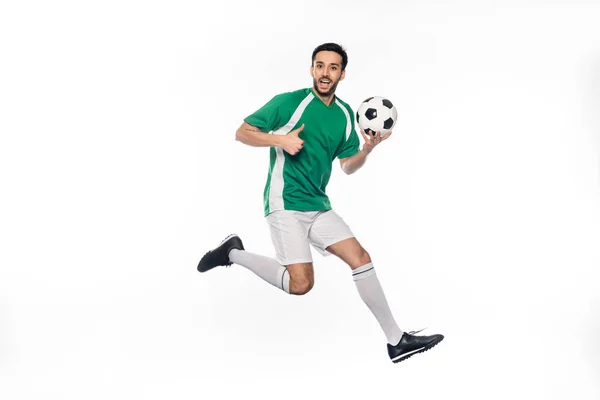Sorprendido jugador de fútbol en uniforme saltando mientras sostiene la pelota de fútbol y mostrando el pulgar hacia arriba en blanco - foto de stock
