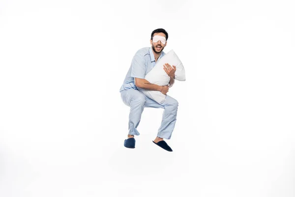 Homme heureux en pyjama et masque de sommeil lévitant avec oreiller sur blanc — Photo de stock