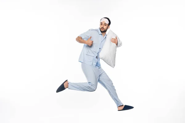 Bearded man in pajamas and sleeping mask levitating with pillow while showing thumb up on white — Stock Photo