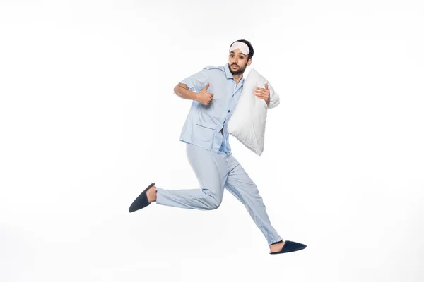 Homme barbu en pyjama et masque de sommeil lévitant avec oreiller tout en montrant pouce vers le haut sur blanc — Photo de stock