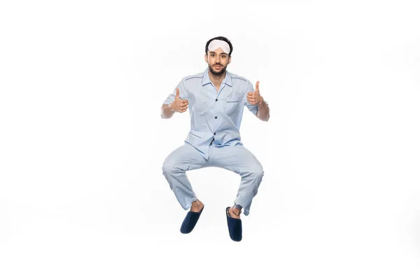 Happy bearded man in pajamas and sleeping mask levitating while showing thumbs up on white — Stock Photo