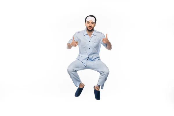 Hombre barbudo feliz en pijama y máscara de dormir levitando mientras muestra los pulgares hacia arriba en blanco - foto de stock